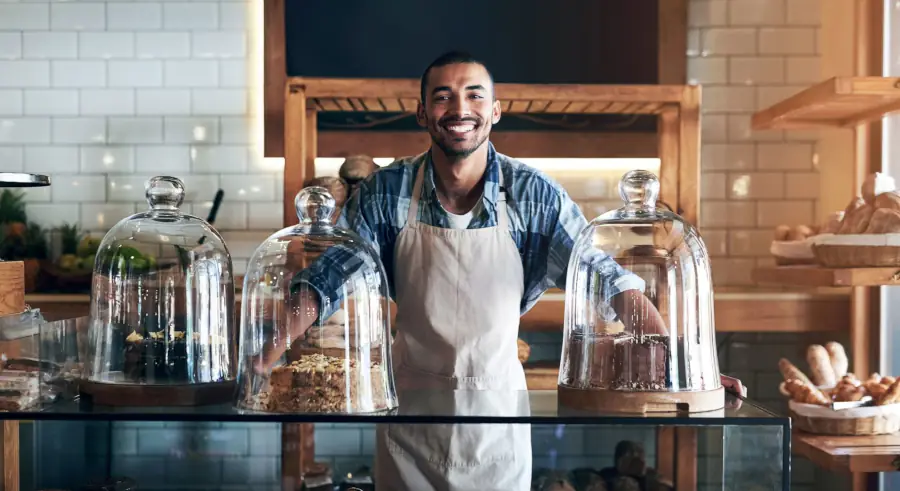 Bakery employee standing behind glass counter with pastries under display domes in welcoming café setting.
